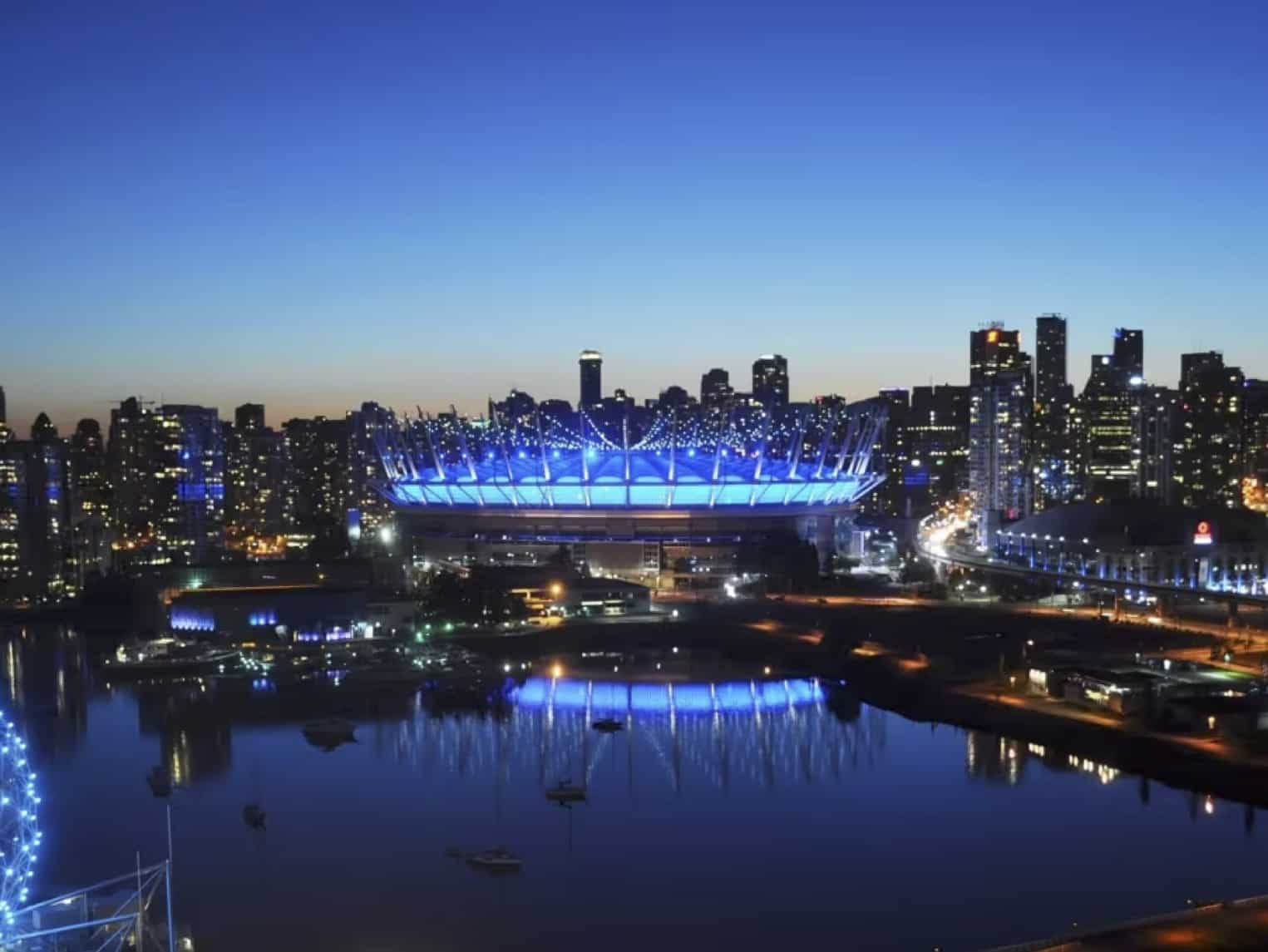 Night cityscape at dusk with a blue-lit stadium reflecting in calm water near a harbor