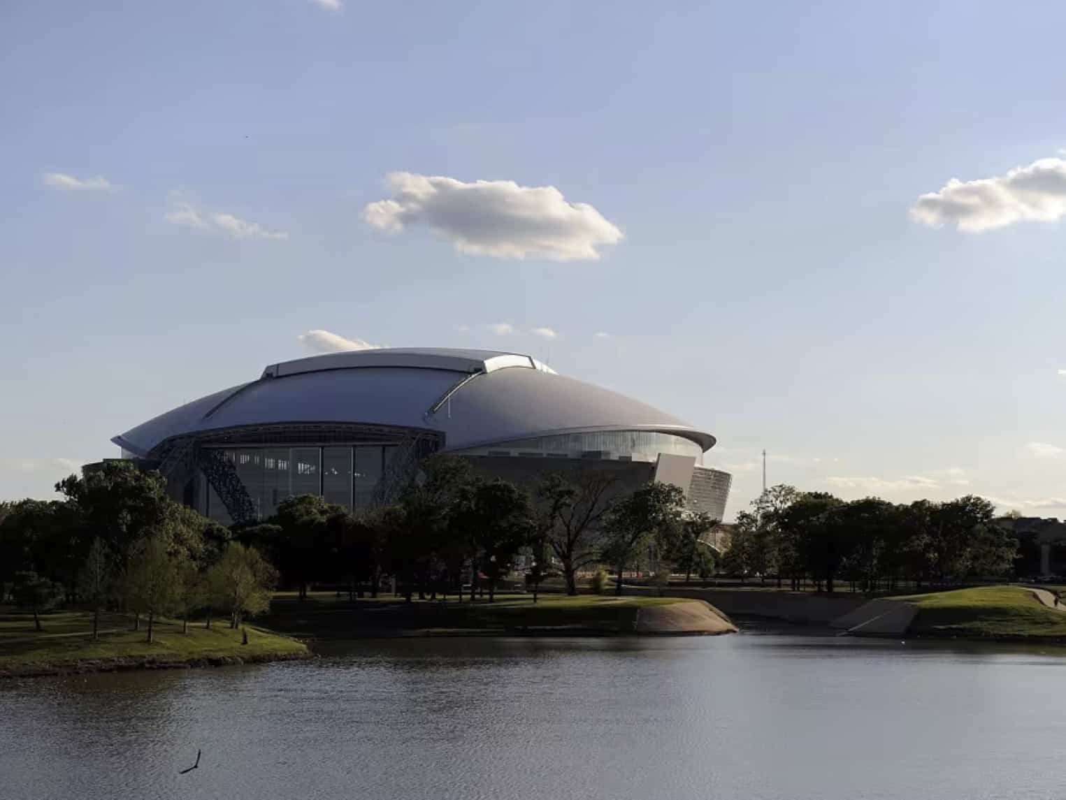 Modern, curved-roof stadium surrounded by trees by a lake on a clear day; blue sky with a few clouds in the background.