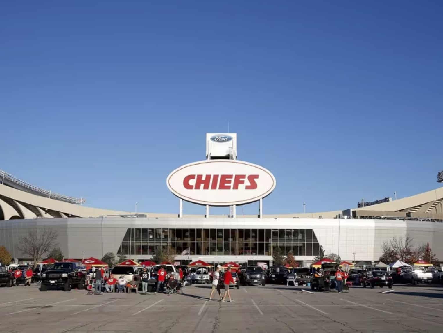 Exterior view of a Chiefs stadium with a large oval 'CHIEFS' sign atop the building, and fans tailgating in the parking lot below.