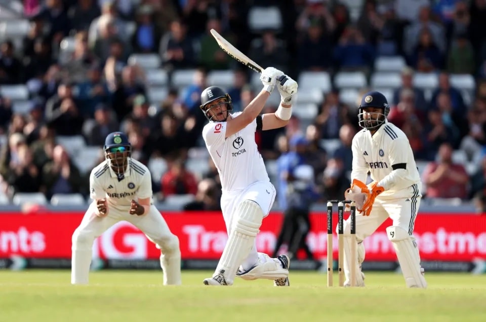 Cricketer in white swings his bat during a shot as two fielders and a cheering crowd watch.